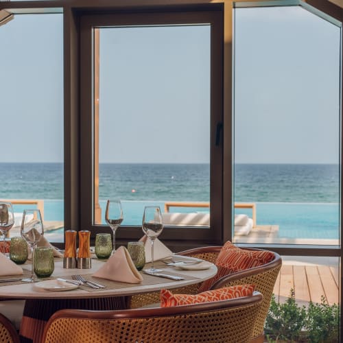 Eleganter gedeckter Esstisch mit gefalteten Servietten, Weingläsern und grünem Glas in einer Strandvilla mit Blick auf einen Pool und das Meer durch große Fenster.