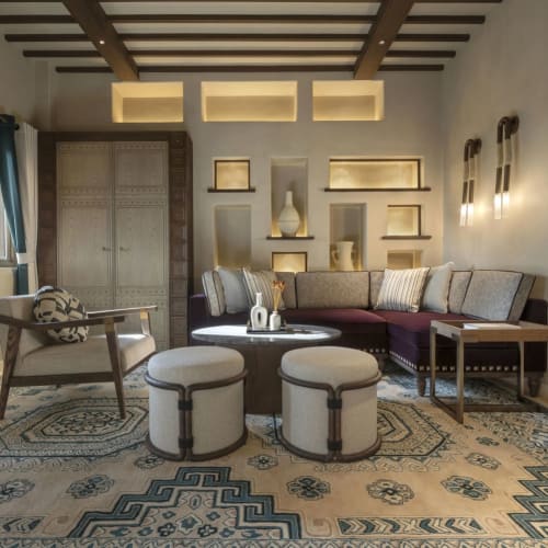 Elegantes Wohnzimmer mit Decke aus Holzbalken, gemustertem Teppich, Ecksofa, zwei runde Ottomane und Wandnischen mit Dekorationsgegenständen.