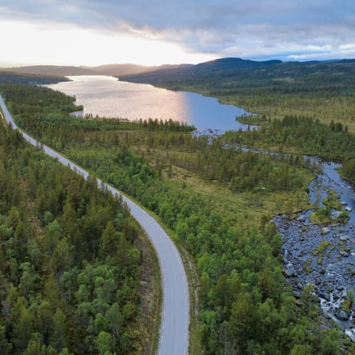 A scenic aerial view of a winding road alongside a river and a lake in a lush green landscape in Norway.