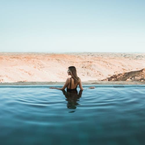 Eine Frau in einem schwarzen Badeanzug steht in einem Infinity-Pool mit Wüstenhügeln im Hintergrund unter klarem Himmel.