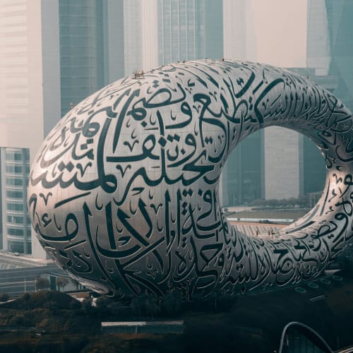 Ein futuristisches, großes, schlingenförmiges Gebäude mit arabischer Kalligraphie auf seiner Oberfläche in einer Stadt mit modernen Wolkenkratzern.
