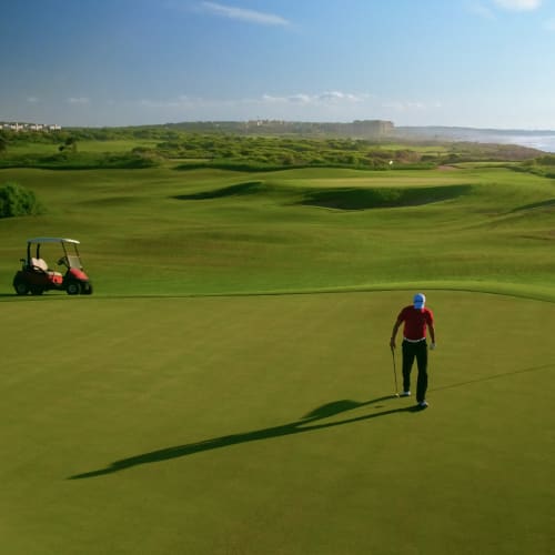 Golfer walking on a green golf course at Mazagan Beach Golf Resort with a golf cart nearby and the ocean in the background under a clear blue sky.