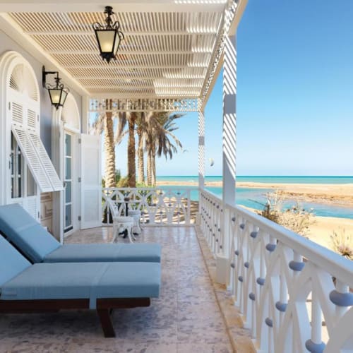Balkon mit blauen Liegestühlen und weißem Holzgeländer mit Blick auf einen Sandstrand, Palmen und ein ruhiges Meer unter klarem blauem Himmel.