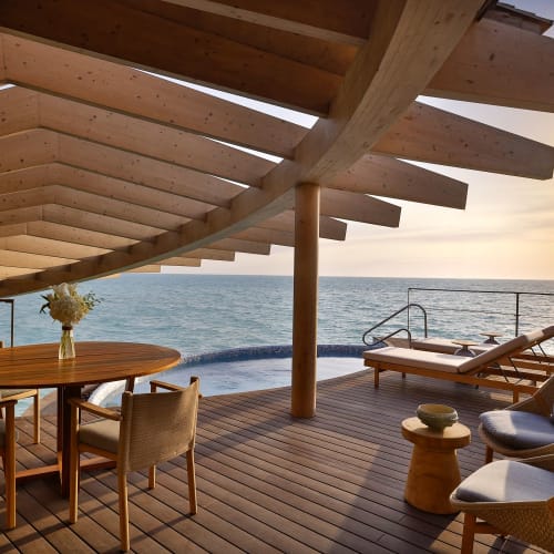 Ein luxuriöser Meeresblick-Balkon mit Holzfußboden, Sitzgelegenheiten im Freien, einem Esstisch mit Blumenvasen und Liegestühlen mit Blick auf den Ozean bei Sonnenuntergang.