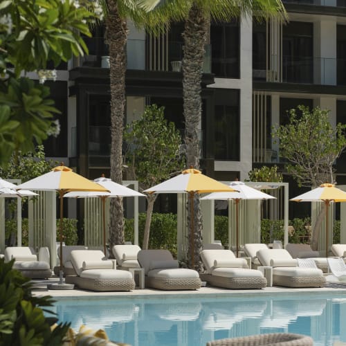 Luxuriöse Poolseite mit weißen Liegen, gelb-weißen Sonnenschirmen und hohen Palmen vor einem modernen Gebäude.