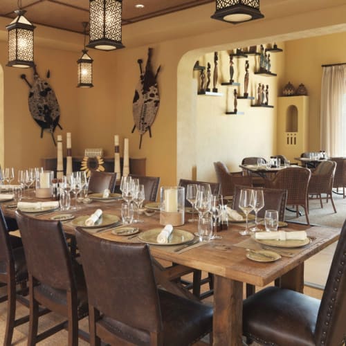 Ein geräumiger Essbereich in einem Resort mit einem langen Holztisch, gedeckt mit Tellern, Gläsern und Servietten, umgeben von Ledersesseln, großen Fenstern und afrikanischer Wanddekoration mit Schilden und Skulpturen.