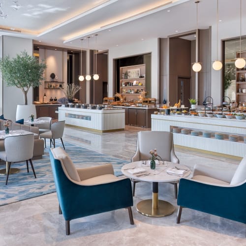 Luxuriöses Hotelrestaurant mit modernem Dekor, weißen und blauen Stühlen, einem Marmortisch und einem Frühstücksbuffet im Hintergrund.