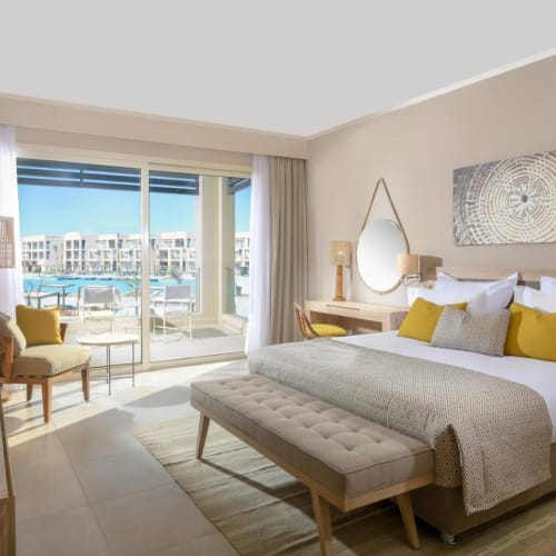 A luxurious hotel room with a large bed, yellow and white pillows, and a balcony with a view of the resort and water.