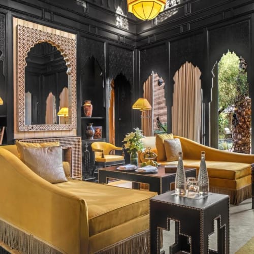 Luxurious living room with yellow velvet sofas, ornate black walls, and large arched windows revealing greenery outside.