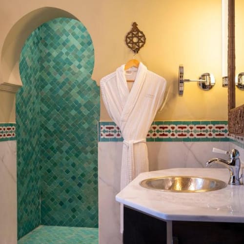 Ein Badezimmer mit einem kleinen Duschbereich in grünem Mosaikfliesen, einem weißen Bademantel, der an der Wand hängt, und einem Waschbecken mit goldener Schale.