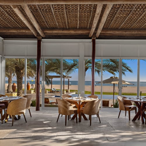 Innenbereich eines Speiseraums mit runden Holztischen und Korbsesseln vor großen Glasfenstern mit Blick auf einen Strand mit Palmen und Sonnenschirmen unter blauem Himmel.
