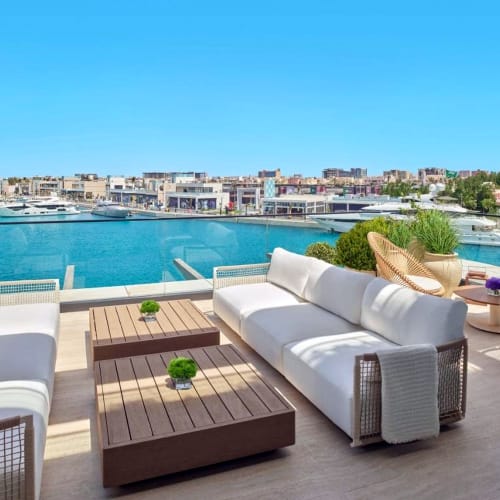 Luxuriöse Outdoor-Lounge mit weißen Sofas und Holztischen mit Blick auf den Jeddah-Yachthafen und die Skyline.