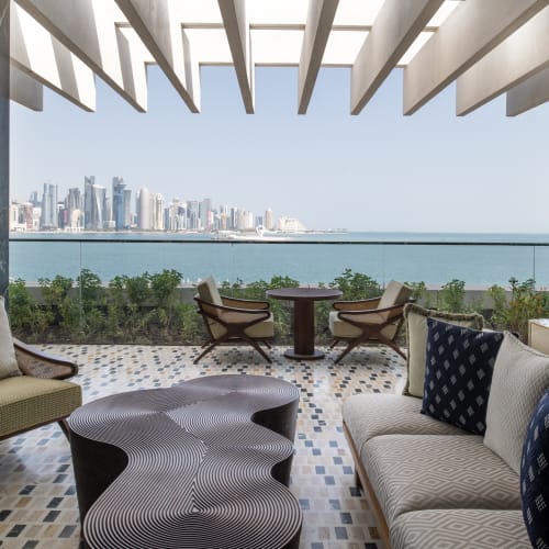 Moderner Außenloungebereich im The Ned Doha mit stilvollen Stühlen, einem Sofa und einem gemusterten Couchtisch mit Blick auf die Skyline von Doha über das Wasser.