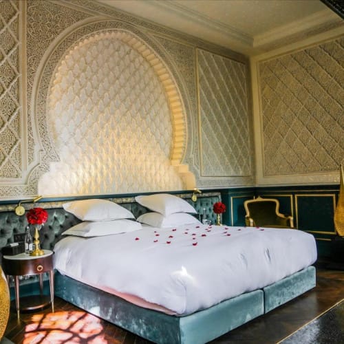 Luxuriöses Schlafzimmer in einer Riad mit einer kunstvollen, gemusterten Wandnische über einem großen Bett mit weißen Bettlaken und Kissen, flankiert von goldenen dekorativen Vasen und kleinen Beistelltischen mit roten Blumen.