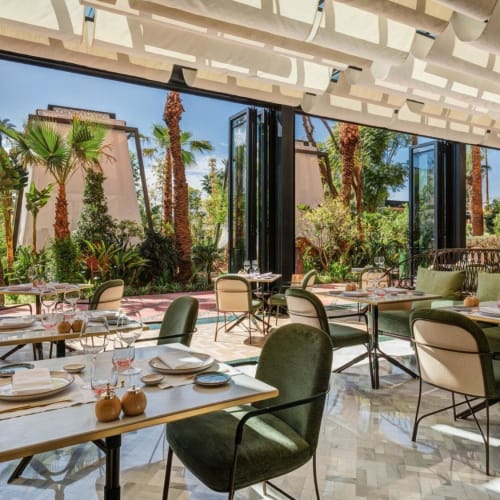  Elegantes Restaurantinnenraum mit großen offenen Glastüren, die einen grünen Garten mit Palmen draußen zeigen.