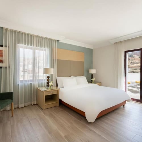 A spacious hotel room with a large bed, two bedside lamps, a sitting area, and a balcony overlooking a scenic landscape.