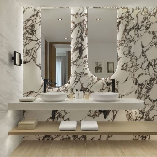 Ein modernes Badezimmer mit einer großen Marmorwand mit weißen und braunen Adern, zwei stilvollen ovalen Spiegeln und einer langen weißen Ablage mit zwei Waschbecken und schwarzen Armaturen.