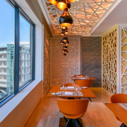 Ein modernes asiatisches Restaurant mit Holztischen und orangefarbenen Stühlen, dekoriert mit stilvoller Beleuchtung und geometrischen Wandtrennwänden, neben großen Fenstern mit Blick auf die Stadt.
