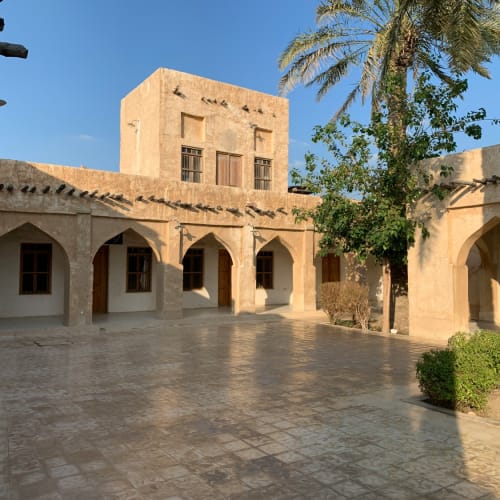 Sunlit courtyard surrounded by traditional Middle Eastern architecture, with arched doorways, palm trees, and a clear blue sky.