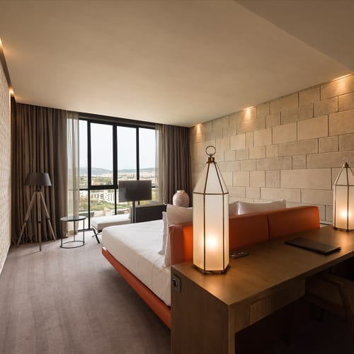 Ein modernes Hotelzimmer mit großem Fenster und Stadtblick, ausgestattet mit einem stylischen Bett mit orangefarbenem Kopfteil, zwei hohen Laternenlampen auf einem Holztisch und strukturierten Wänden.