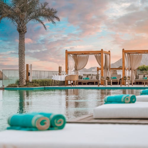 Unendlichkeitspool in einem luxuriösen Strandresort mit weißen Liegen und türkis gerollten Handtüchern, hölzernen Pavillons mit Vorhängen, Palmen und einer malerischen Meeres- und Felseninselkulisse bei Sonnenuntergang.