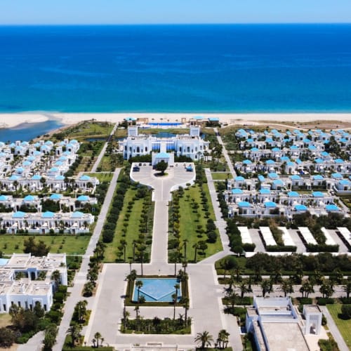 Luftaufnahme des Banyan Tree Tamouda Bay Resorts mit weißen Gebäuden, grünen Rasenflächen und Schwimmbädern, gelegen an einem Sandstrand mit blauem Meer im Hintergrund.