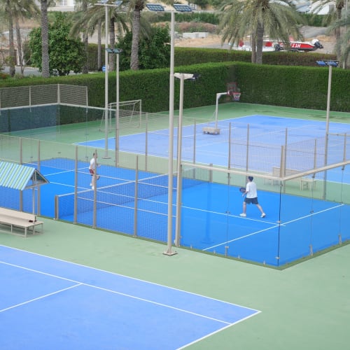 Zwei Personen spielen Padel auf einem blauen Außenplatz, umgeben von Glaszäunen und Palmen in einer sonnigen Umgebung.