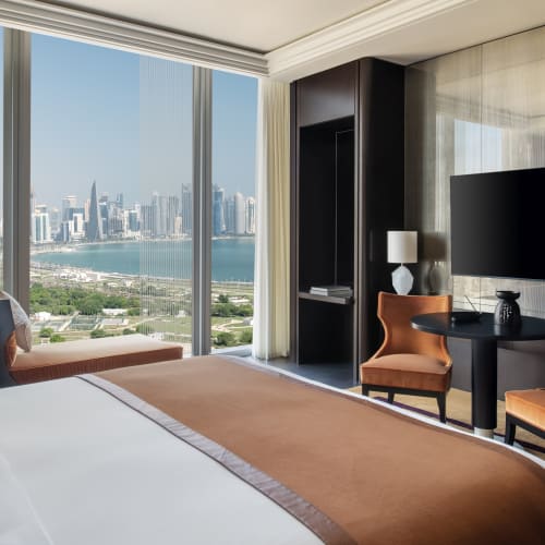 Modernes Hotelzimmer mit großem Bett, zwei orangefarbenen Stühlen, einem Tisch und einem Flachbildfernseher, mit Panoramablick auf die Stadtsilhouette und das Wasser durch bodentiefe Fenster.