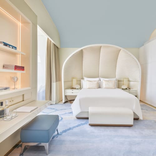 Helles und modernes Hotelzimmer mit einem einzigartig geschwungenen weißen Betthaupt, einem großen Bett mit weißer Bettwäsche, Nachttischen mit Lampen, einer blauen Bank und einem hellen Teppich.