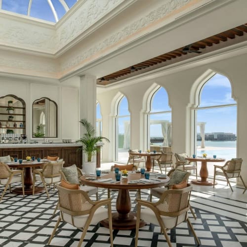 Luxuriöse Lounge mit großen bogenförmigen Fenstern mit Blick auf das Meer, mit runden Tischen, eleganten Stühlen und einem Musterboden.