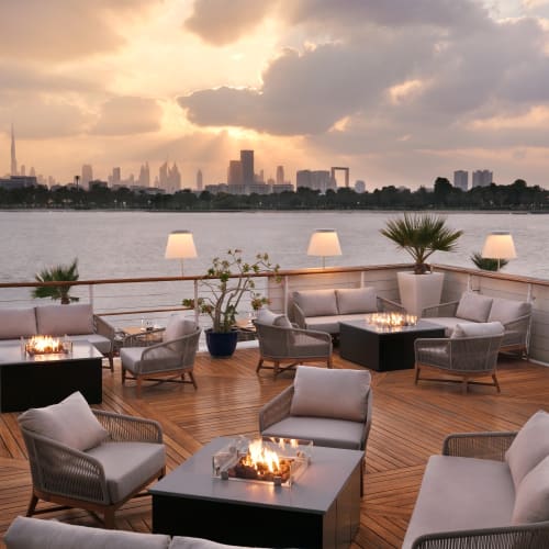 Außenlounge im Park Hyatt Dubai mit modernen Sitzmöbeln, Feuerschalen und Stehlampen auf einem Holzdeck mit Blick auf einen Fluss und die Stadtsilhouette unter einem bewölkten Sonnenuntergangshimmel.