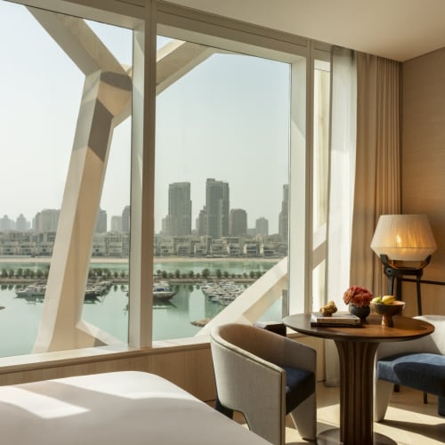 The hotel room features a round table with chairs, set near large windows overlooking a river and city skyline.