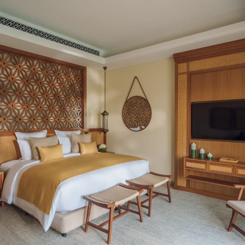 Luxuriöses Schlafzimmer in den Naäma Beach Villas and Spa mit großem Bett in goldbraunen Tönen, dekorativem Holzpaneel an der Wand, zwei gewebten Hockern am Fußende und einer an der Wand montierten Flachbildfernseher.