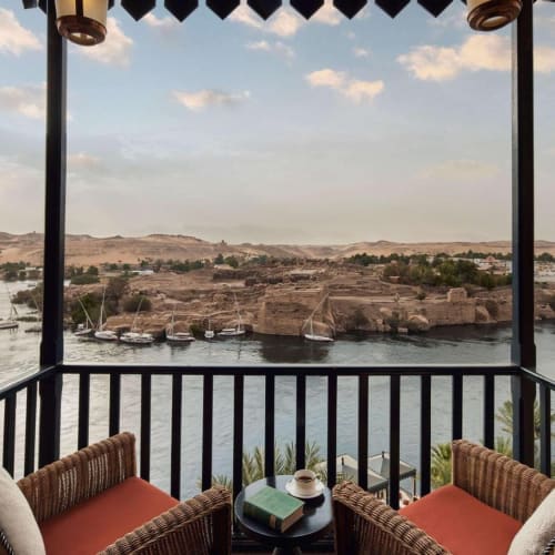 Ein Balkon mit Blick auf den Nil mit zwei Rattanstühlen und einem kleinen Tisch, eingerahmt von einer dunklen Struktur, mit Booten auf dem Wasser und einer Wüstenlandschaft im Hintergrund.