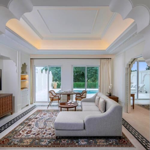 Luxus-Inneneinblick in ein Wohnzimmer mit weißen Wänden, einem weißen Sofa, einem Flachbildfernseher, einem Holztv-Schrank, einem bunten Teppich und großen Schiebetüren, die zu einem Poolbereich führen.