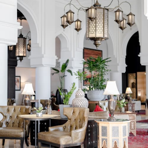 Luxurious hotel lobby with elegant beige armchairs, ornate lamps, and lush green plants under intricate Moroccan-style lanterns.
