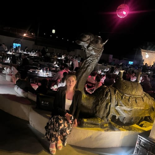Frau sitzt auf einer niedrigen Mauer neben einer großen bronzenen Kamelskulptur in einem Außenbereich mit Tischen zum Essen bei Nacht, viele Menschen sitzen im Hintergrund unter gedämpftem Licht, darüber eine leuchtend pinke Kugel als Lichtquelle.