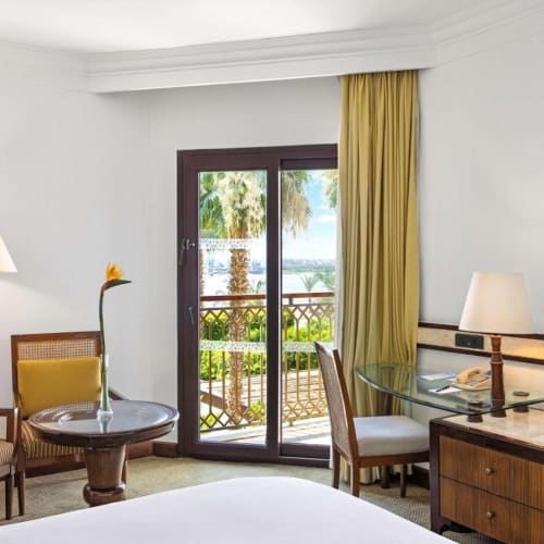 A hotel room sitting area with two chairs, a small round table with a flower, a lamp, and a balcony overlooking palm trees and water.