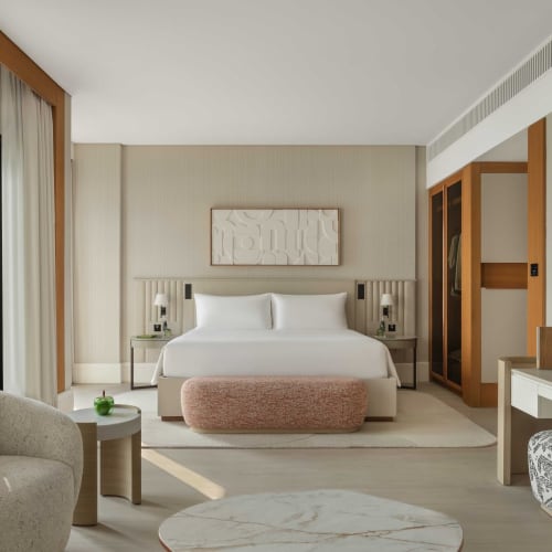 Modern hotel room with a large bed, side tables, a TV, and a sitting area with a sofa in a bright setting.