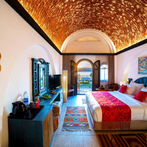 A cozy hotel room with a double bed adorned with red and beige bedding, a wooden media console with a TV, and a rustic arched brick ceiling. The room opens to a balcony overlooking a scenic waterfront view under a bright sky.