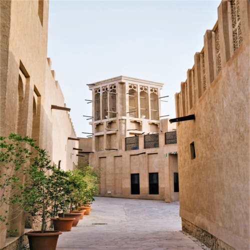 A narrow alleyway in Dubai's Al Fahidi historical district with traditional wind-tower buildings and potted plants lining the path.