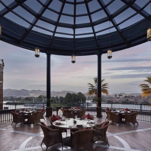 Ein Restaurant mit einem gewölbten Glasdach und runden Tischen zum Essen, mit Blick auf Palmen und die Stadt bei Sonnenuntergang.