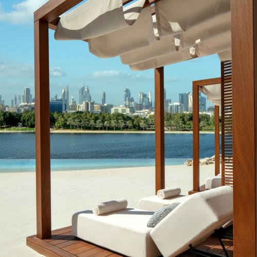 Liegestühle unter einem Baldachin auf einer Terrasse mit Blick auf einen Fluss und die Skyline von Dubai.