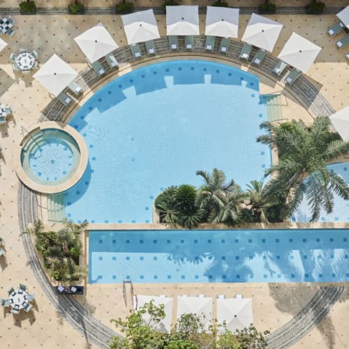 Luftaufnahme eines Hotelpoolbereichs mit einem großen geschwungenen Schwimmbecken, einem kleineren runden Becken und einem rechteckigen Becken, umgeben von weißen Sonnenschirmen, Liegestühlen und Palmen.