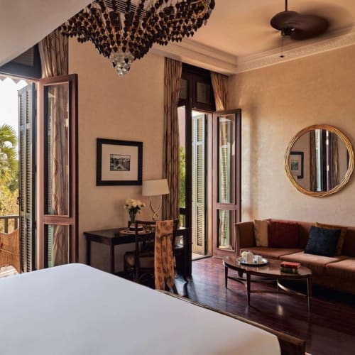 Ein luxuriöses Hotelzimmer mit offenen Balkonstellen, die Palmen draußen zeigen, ein großes Bett, ein Sitzbereich mit einem Sofa und elegante Dekoration.