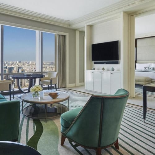 Luxurious living room with large window overlooking a city skyline, featuring teal and cream furniture, a flat-screen TV, and modern decor.
