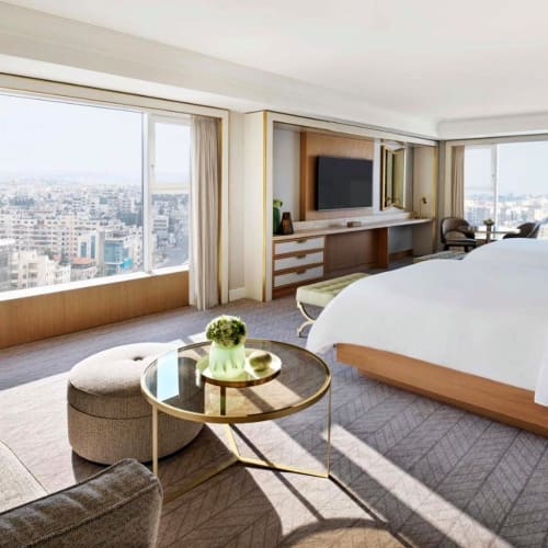 Ein luxuriöses Hotellzimmer mit großen Fenstern, die eine Stadtansicht zeigen, einem Kingsize-Bett, einem Sofa, einem runden Couchtisch und einem Flachbildfernseher.