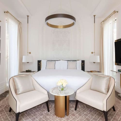 Ein elegantes Schlafzimmer mit einem großen, weißen Bett, flankiert von zwei Nachttischen und Lampen, sowie zwei sitzenden Sessel um einen kleinen, runden Tisch. Der Raum ist hell erleuchtet mit Vorhängen vor den Fenstern und moderner Dekoration.