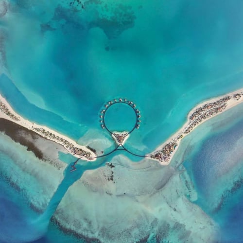 Luftaufnahme eines luxuriösen Resorts mit kreisförmigen Wasserbungalows, die durch eine Brücke verbunden sind, umgeben von türkisfarbenem Wasser und Sandstränden.
