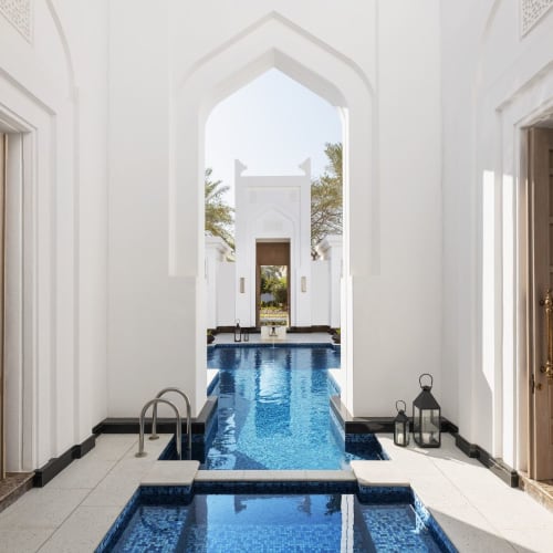 Luxuriöse Innenpool-Area mit weißen Wänden und Blick nach draußen durch eine gewölbte Tür im Raffles Al Areen Palace Bahrain.
